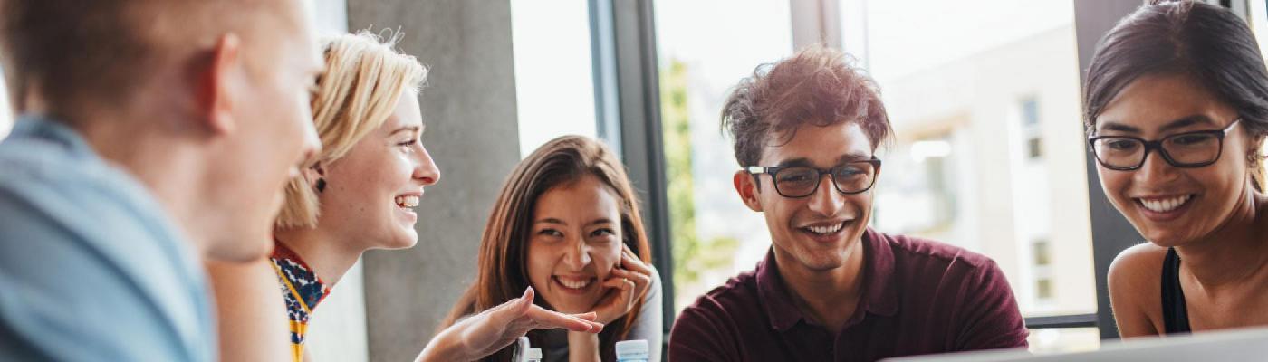 5 Students sitting at a round table looking at a laptop on the table, while talking