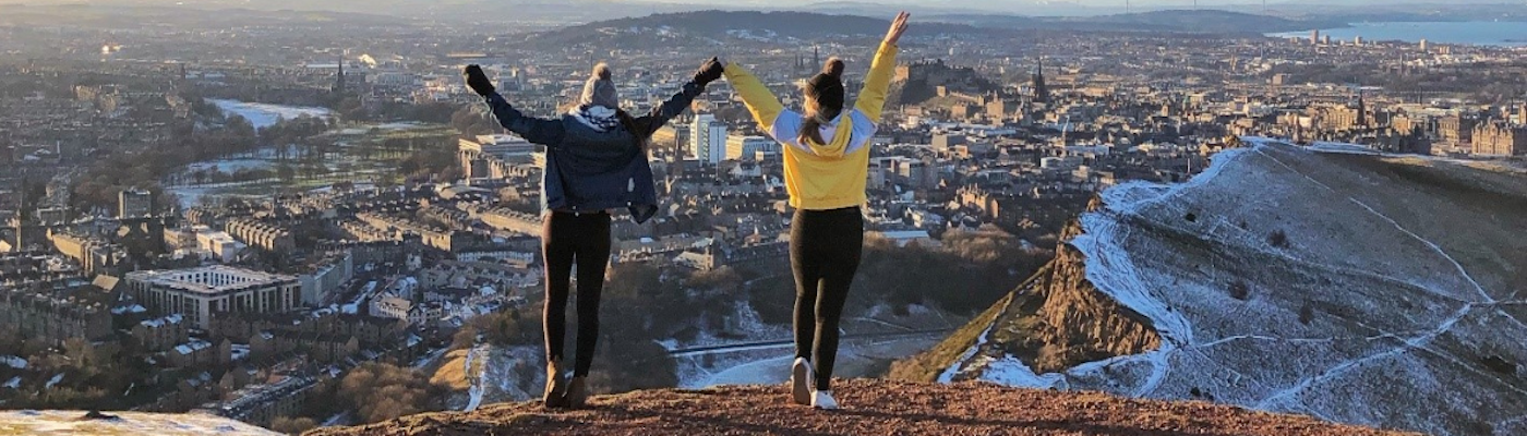 Two students standing on top of a cliff, looking over a city. 