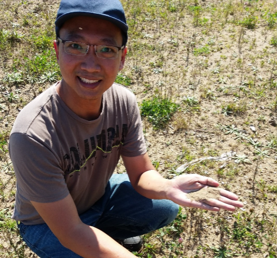 grad student Kai Chung kneeling in field with dirt in palm of hand smiling at camera