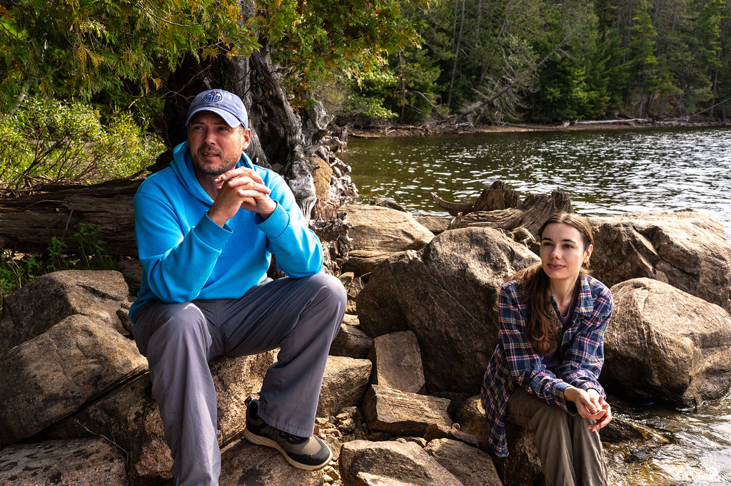 2 students sitting on rocks by the water listening to someone talk
