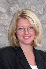 Lady with blonde hair and glasses standing against a rock/concrete wall in business attire and smiling at the camera