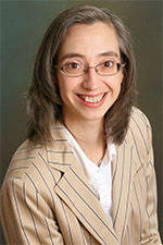 Lady with cream suit and glasses against a muddy green background, smiling at the camera