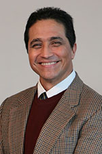 Man with short black hair in a suit and tie smiling at the camera 
