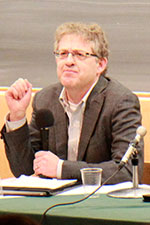 Professor sitting at a desk with a microphone and a blackboard behind him.