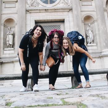 Three girls wearing big backpacks smiling in front of a building in Europe