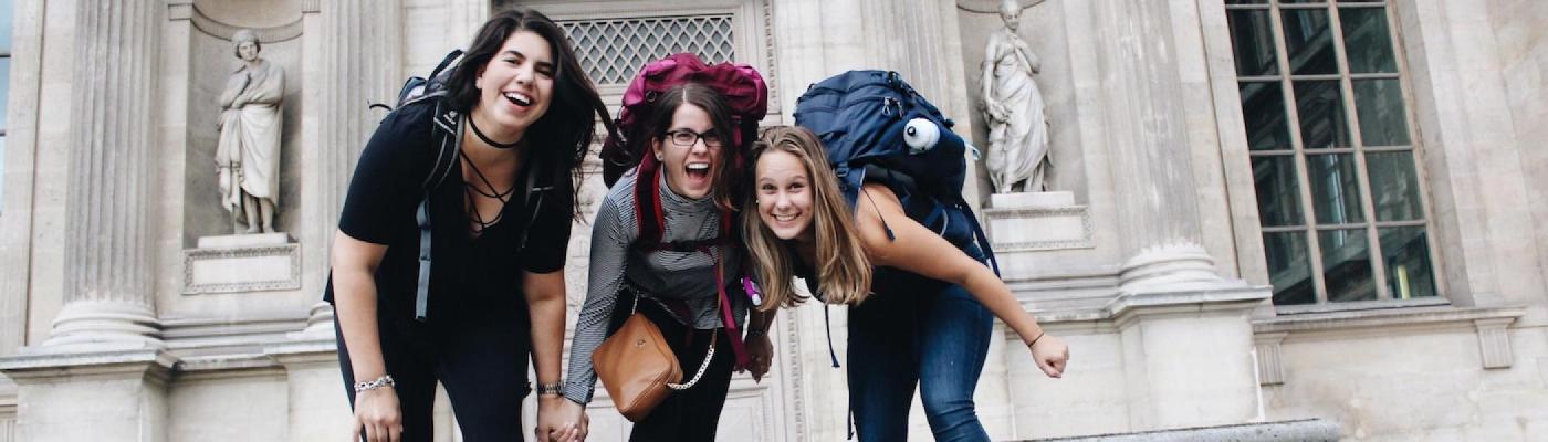 Three girls wearing big backpacks smiling in front of a building in Europe
