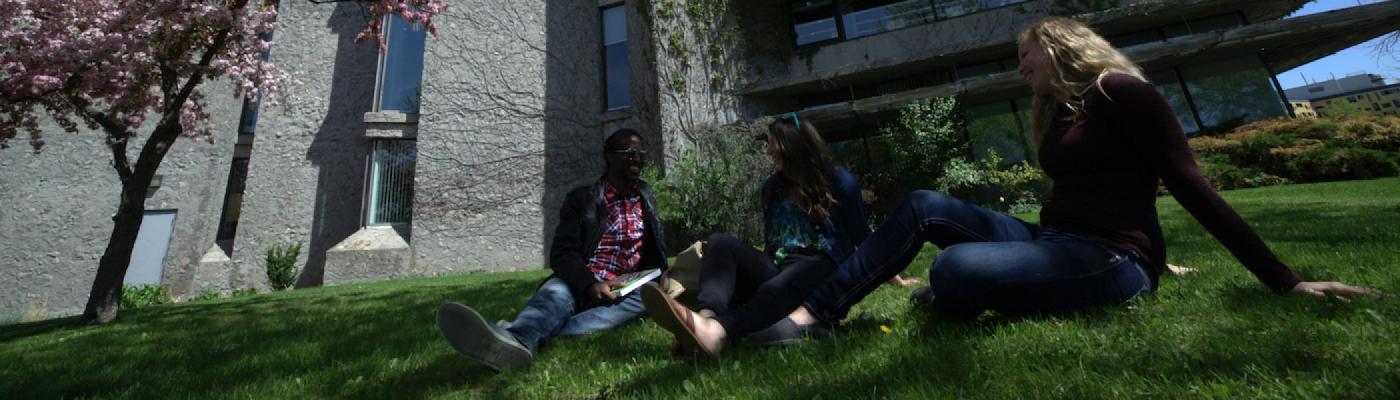 Three students sitting outside on the grass in front of Bata Library smiling