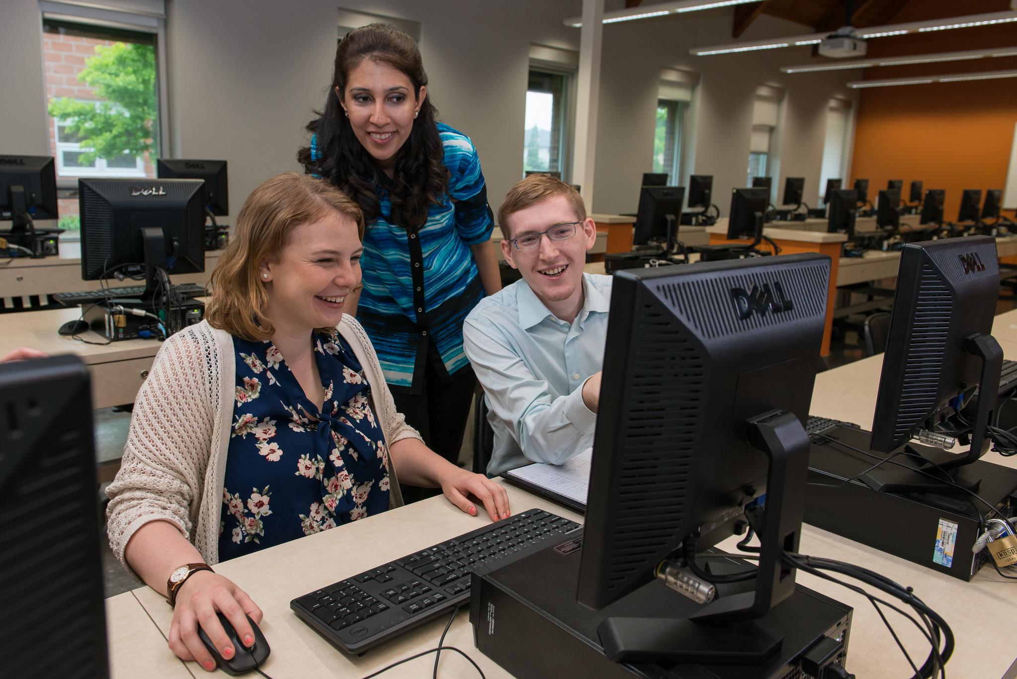 Two students facing a computer, smiling, with teacher leaning in from behind