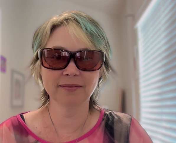 A white woman with short hair with brown, blonde and teal streaks smiles and wears large dark glasses, the kind that protect eyes from light. She wears a pink top in a bright room.