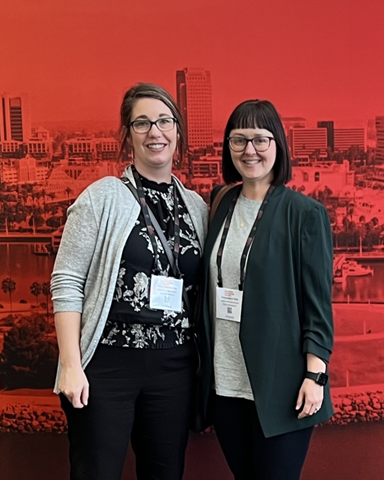 "Holly Bates and Sarah West standing in front of a red tinted cityscape backdrop"