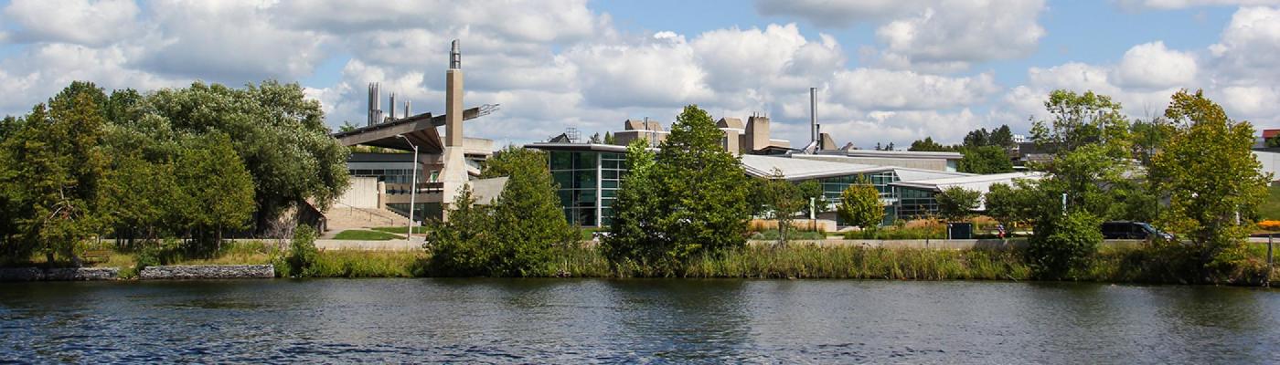 Exterior view of the Science complex from across the Otonabee river on a bright summer's day with blue sky and blue water